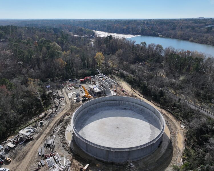 Circular concrete structure under construction, near trees, river, equipment.