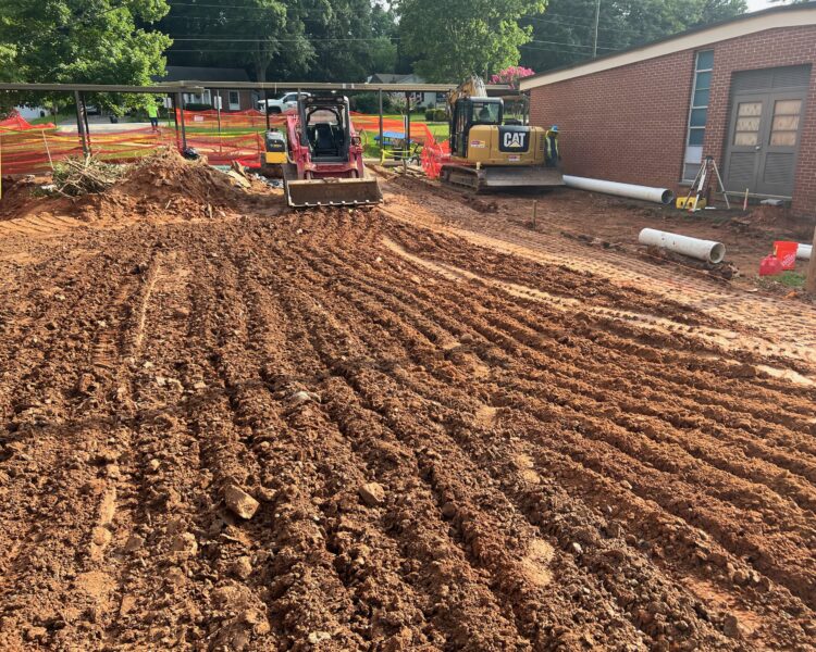 Two excavators on dirt, tire tracks, orange fencing, building behind.