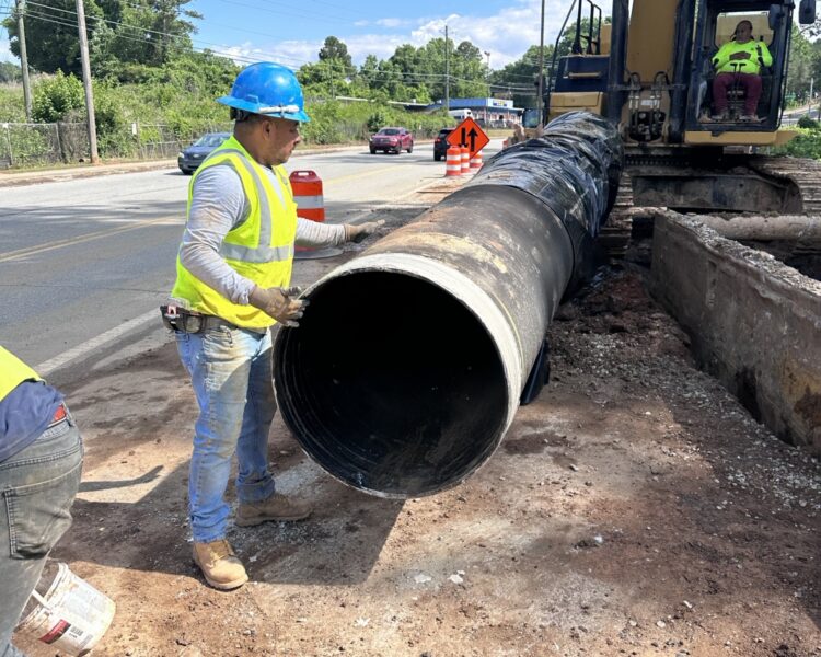 Unos trabajadores equipados con material de seguridad guían una tubería que está levantando una excavadora.