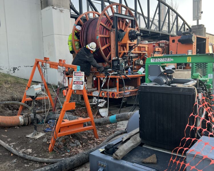 Los trabajadores manejan equipos de cableado junto a un puente, entre maquinaria y barreras de seguridad.
