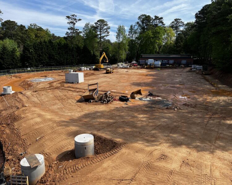 Construction site with machinery, materials, dirt, trees, and building.