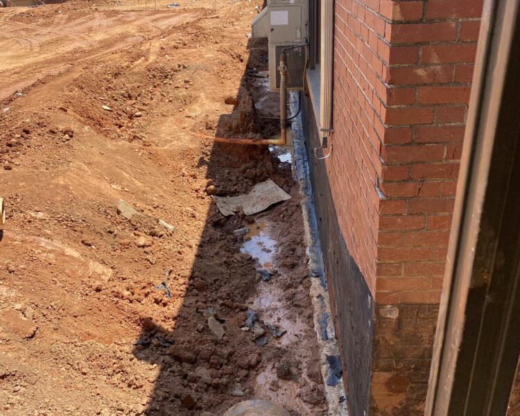Trench with pipe beside brick building; electrical panels on wall.