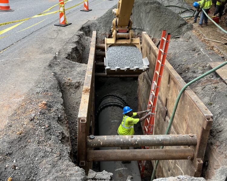 Unos trabajadores instalan una tubería de hormigón en una zanja, cerca de unos conos y una excavadora.