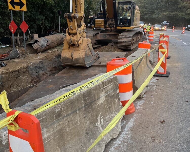Obra en la carretera con una excavadora, conos, vallas y cinta de señalización.