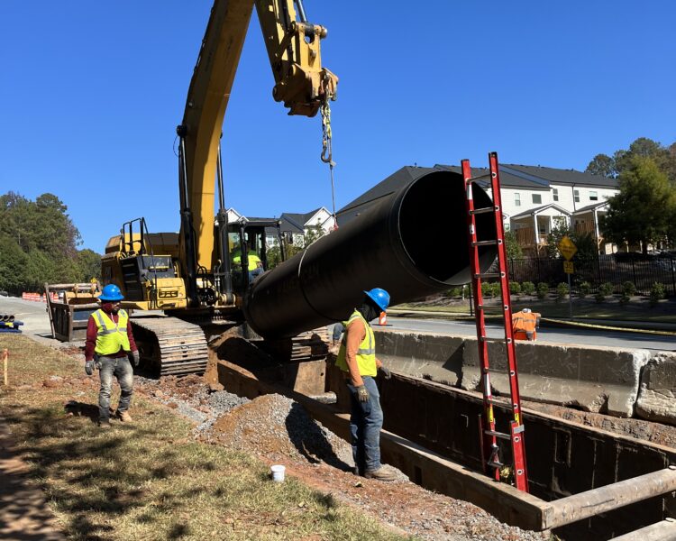 Unos trabajadores y una excavadora bajan un tubo negro a la zanja situada al borde de la carretera.