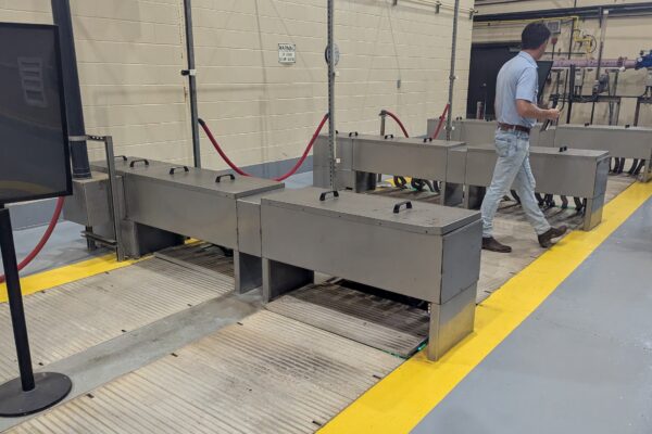 Planta industrial con maquinaria metálica y dos personas junto a una línea amarilla.