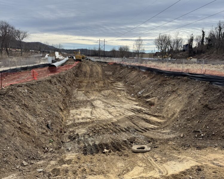 Wide trench at construction site with machinery, cloudy sky.