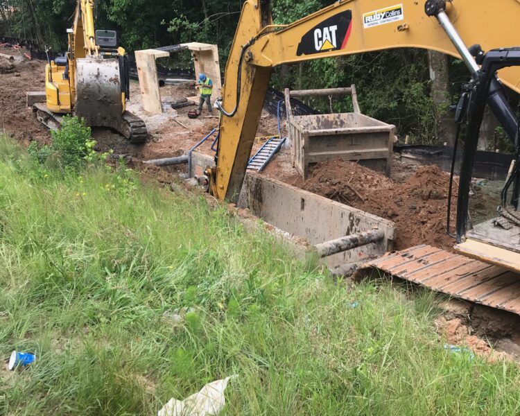 Two excavators operate on mud near trees and trench barriers.