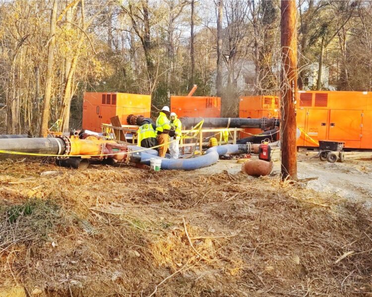 Workers in safety gear by hoses and orange equipment outdoors.