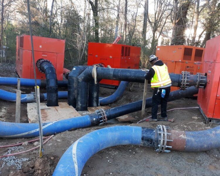 Worker in safety vest checks black and blue pipes outdoors.