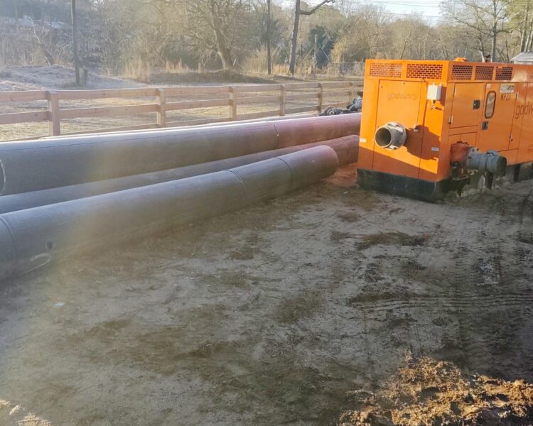 Black pipes beside an orange generator on dirt near a fence.