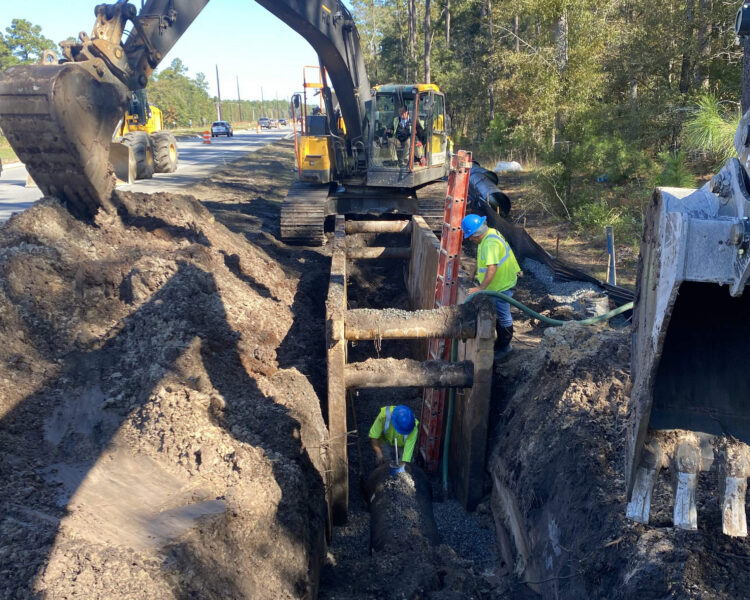 Workers use heavy machinery by a road as one installs pipe underground.