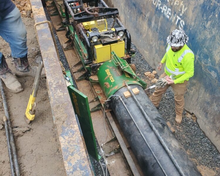 Un trabajador dentro de una caja de zanja utiliza maquinaria para instalar tuberías subterráneas.