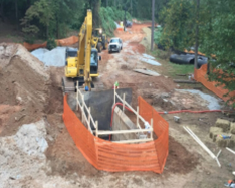 Excavator, workers, and materials at wooded construction site.