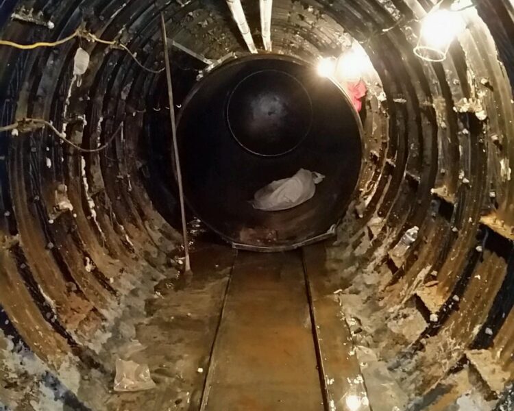 Narrow metal tunnel with pipes, floor water, light, dark pipe.
