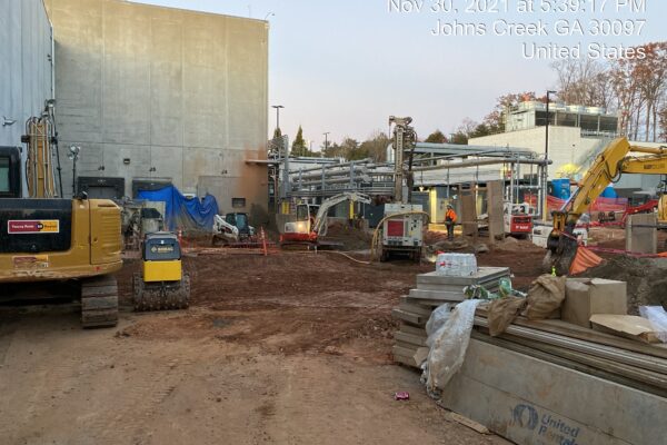 Obreros y máquinas en un terreno sin asfaltar junto a un gran edificio, con superposición de fecha y ubicación.