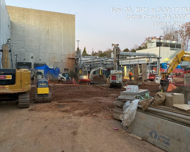Workers and machines on dirt at large building, date/location overlay.