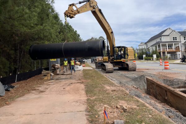 Una excavadora levanta un tubo negro junto a la carretera; hay trabajadores, una zanja y conos de señalización cerca.