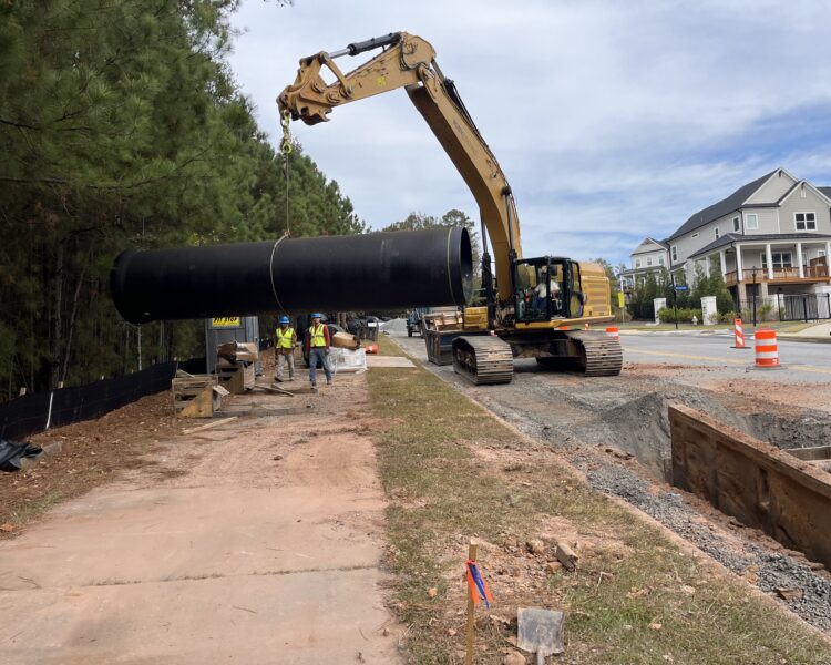 Una excavadora levanta un tubo negro junto a la carretera; hay trabajadores, una zanja y conos de señalización cerca.