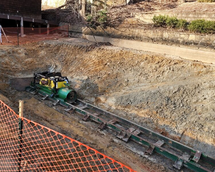 Una pequeña excavadora verde excava una zanja en un terreno sin pavimentar, con una valla detrás.