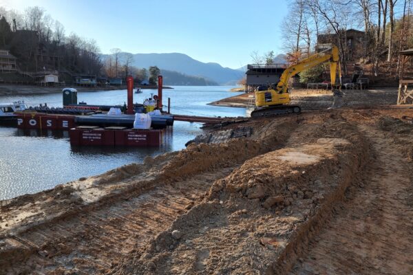 Excavadora amarilla junto a la orilla del lago, con una barcaza y unas casas al fondo.