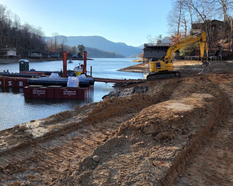 Excavadora amarilla junto a la orilla del lago, con una barcaza y unas casas al fondo.