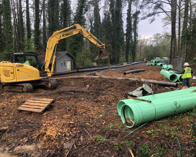 Yellow excavator and worker installing green pipes at forested site.