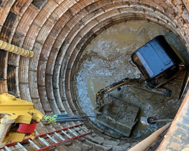 Una pequeña excavadora en el fondo del pozo, rodeada de agua fangosa, con una escalera cerca.