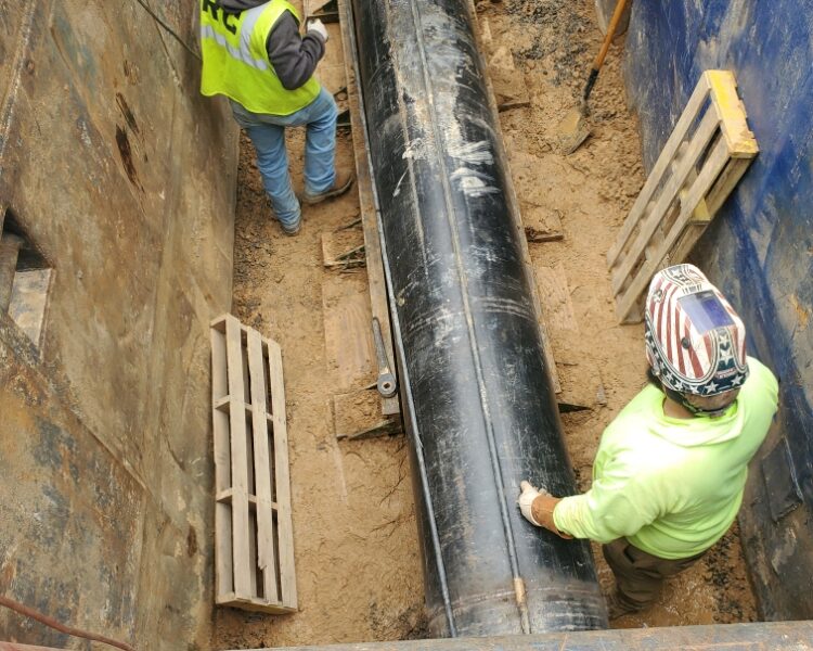 Dos trabajadores inspeccionan una tubería negra en una zanja apuntalada.