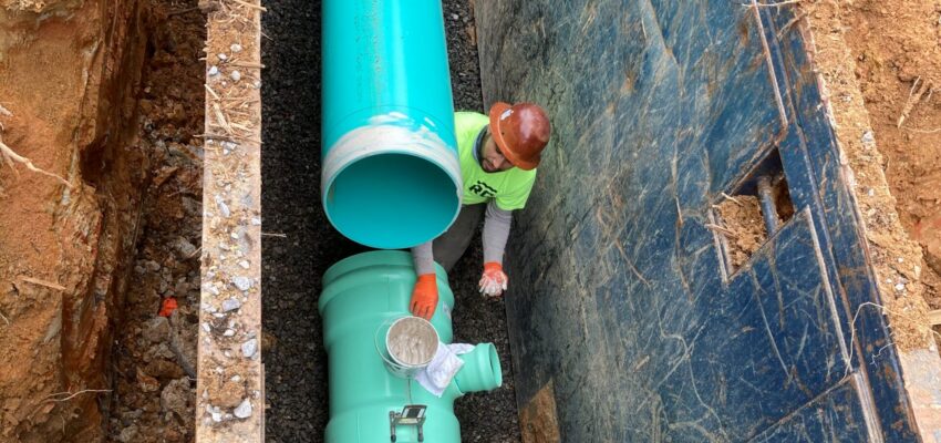 Worker in helmet installs turquoise pipes in narrow trench.