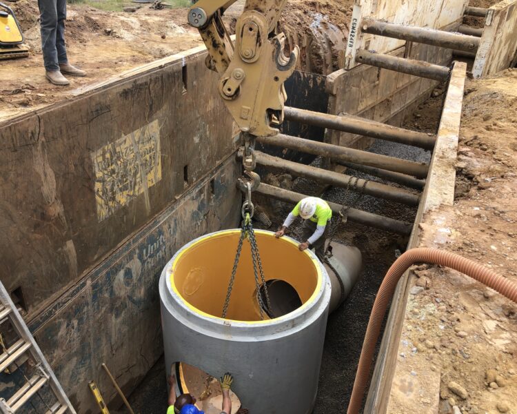 Workers use a crane to lower a concrete pipe into a trench.