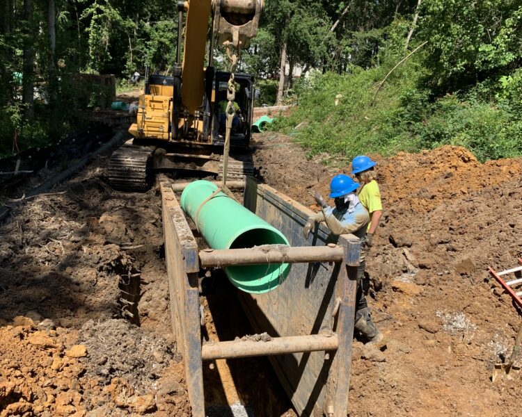 Workers in hard hats guide a green pipe into a trench.