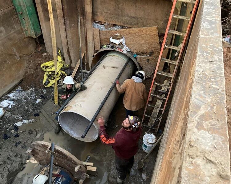 Three workers in muddy trench position large pipe by tools, ladder.