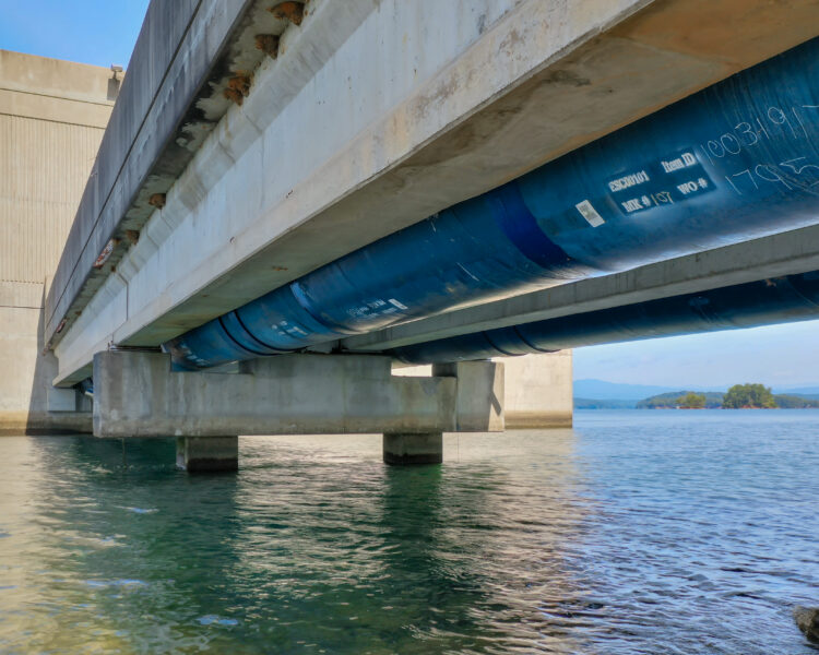Under a concrete bridge with blue pipes over water.