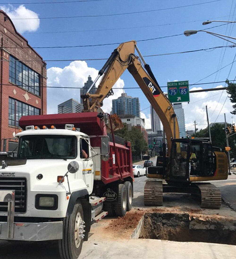 Excavator loads dump truck; workers supervise at city construction site.