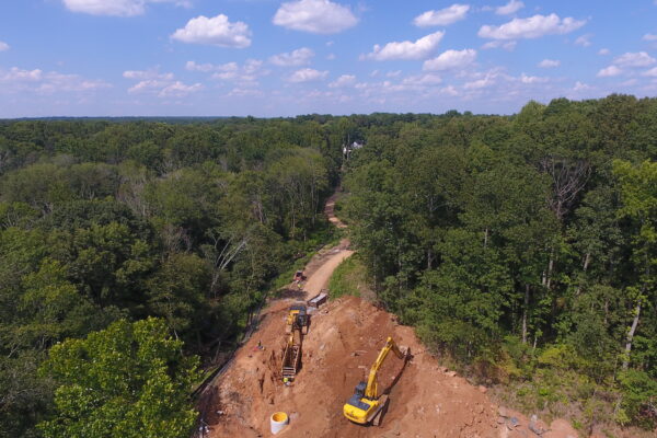 Dos excavadoras en un terreno sin asfaltar, rodeadas de bosque, con el cielo parcialmente nublado.