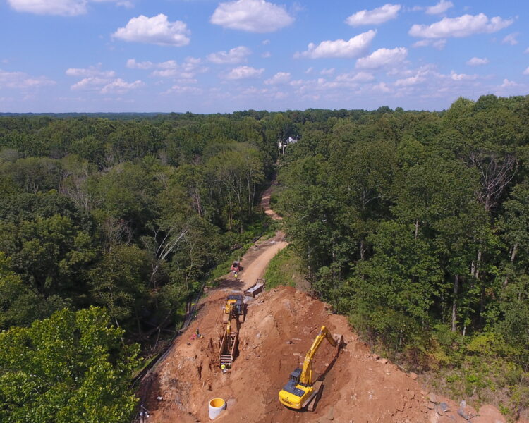 Two excavators on dirt site, forest around, partly cloudy sky.