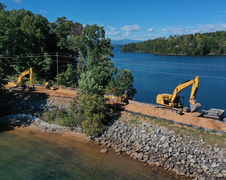 Excavators work on a lakeshore dirt road under blue sky.