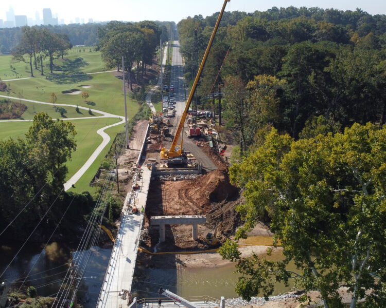 Puente en construcción con una grúa y vehículos cerca del bosque.