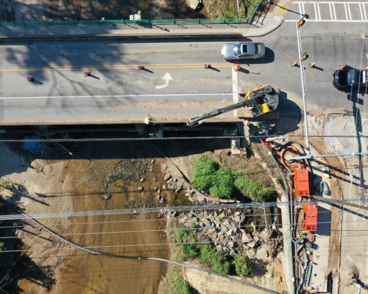 Vista aérea: excavadora, obras en el puente, vehículos y el lecho del río al fondo.