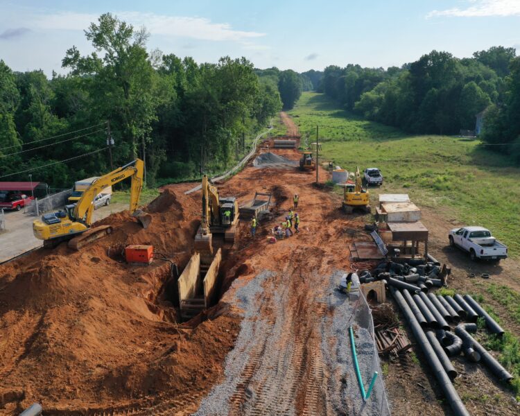 Excavators and workers install large pipes at a dirt site.