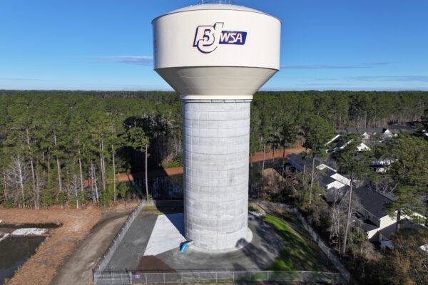 La torre de agua de la BJWSA, con árboles y casas bajo un cielo azul.