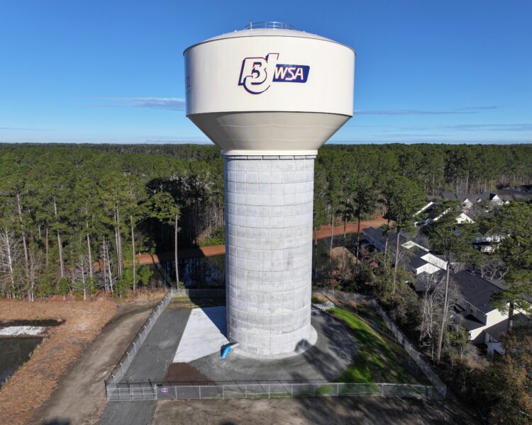 BJWSA water tower with trees and houses beneath blue sky.