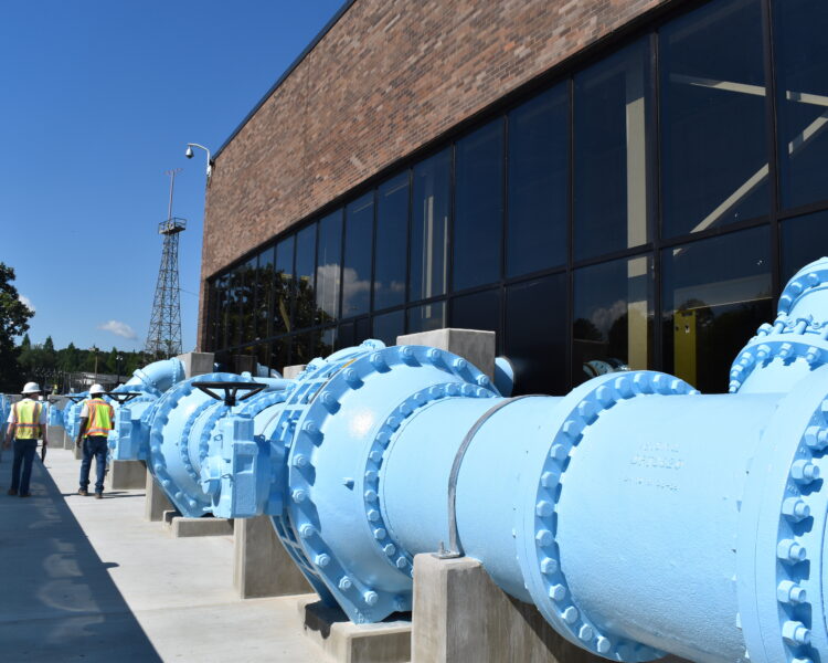 Varias personas inspeccionan unas tuberías industriales azules en el exterior de un edificio de ladrillo.