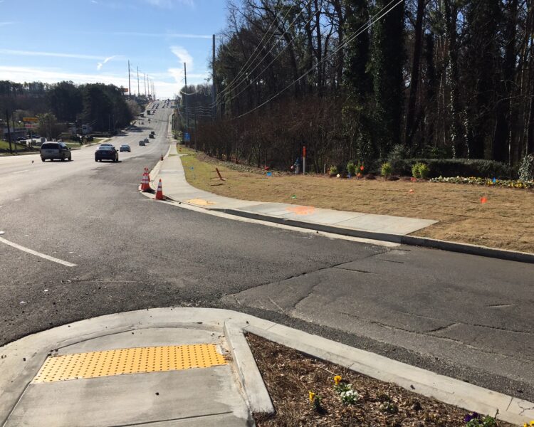 Sidewalk with yellow tactile paving ends at grass beside busy road.