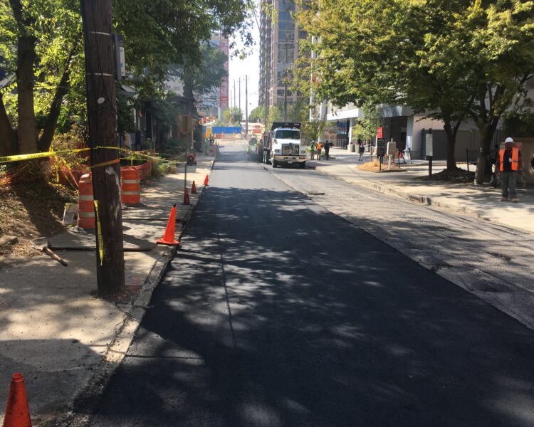 Asfalto recién extendido, conos, obreros, maquinaria y árboles junto a la acera.