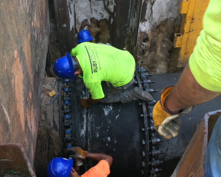Workers in safety gear install a pipe in a narrow trench.