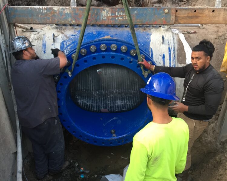 Three workers use a crane to install a large blue pipe.