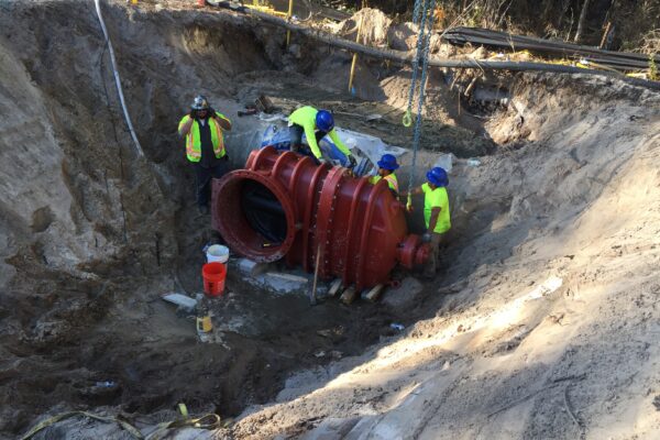 Cuatro trabajadores instalan una gran tubería roja en una excavación profunda.
