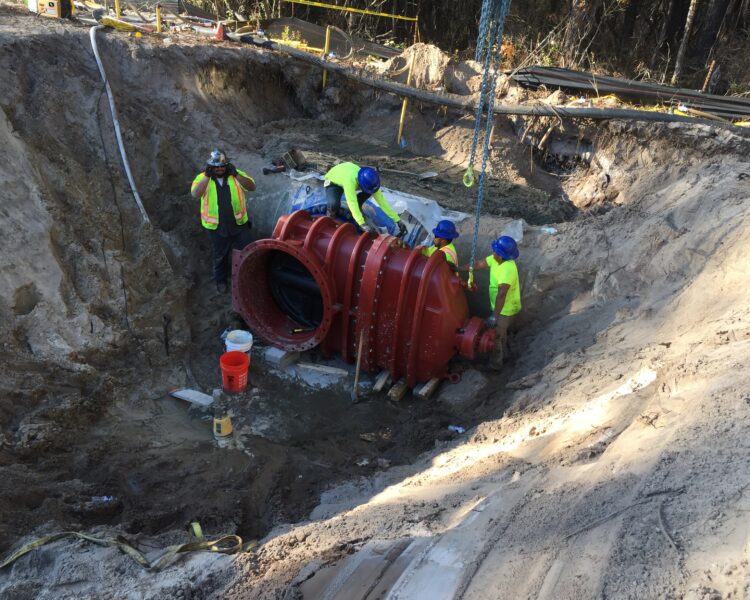 Four workers install a large red pipe in a deep excavation.